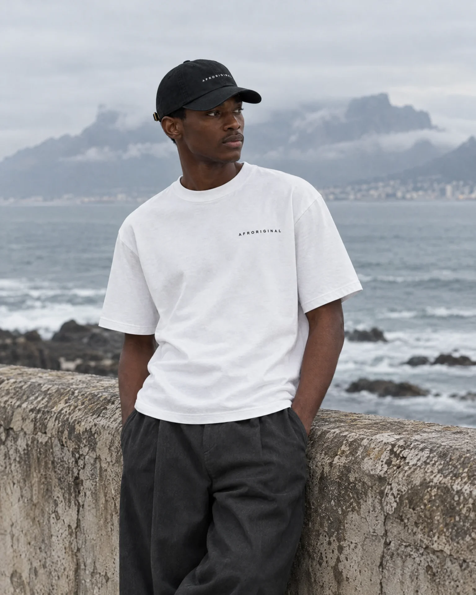 A man in the white Afroriginal wordmark T-shirt and black cap leans on a sea wall with Table Mountain behind him
