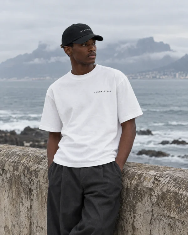 A man in the white Afroriginal wordmark T-shirt and black cap leans on a sea wall with Table Mountain behind him